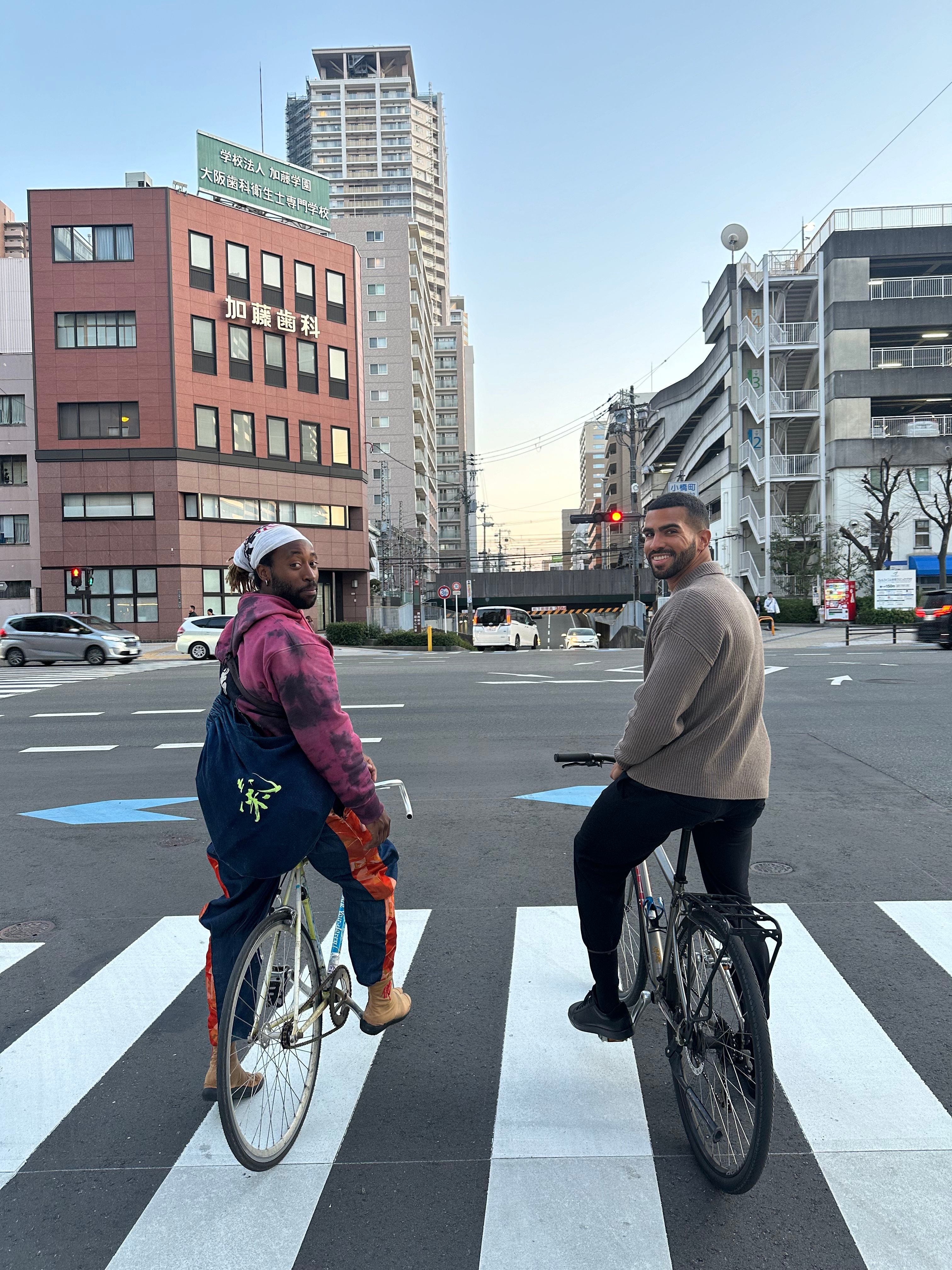 Two people on bicycles crossing a zebra crossing in an urban setting with buildings in the background.